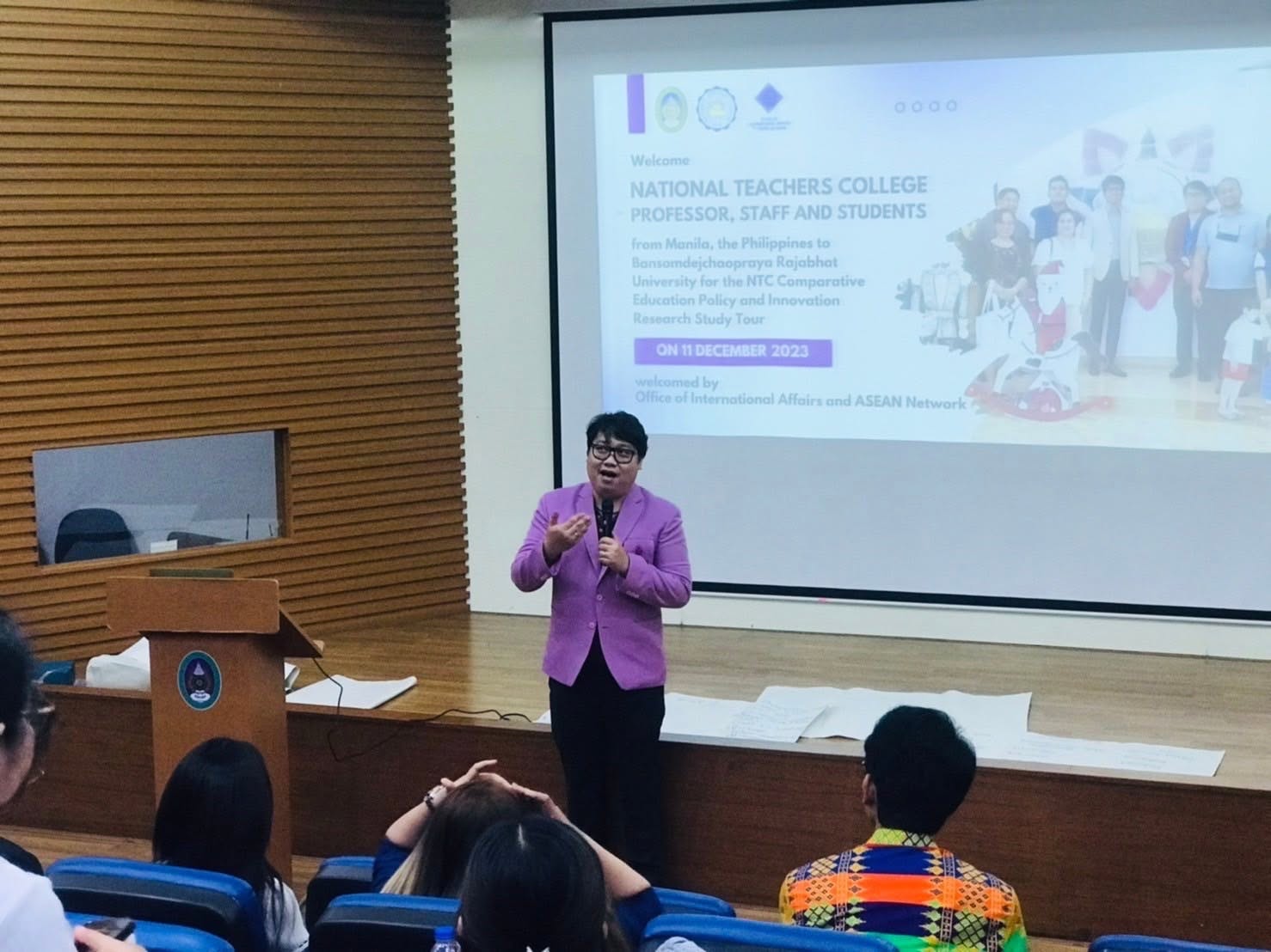 Speaker in a purple suit addressing an audience in a tiered auditorium